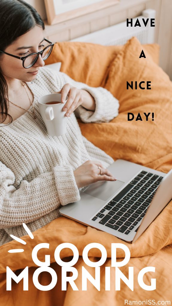 In the photo, a girl wearing a white sweater and glasses is sitting on her yellow bed, using a green tea cup in one hand and using the laptop in the other hand.