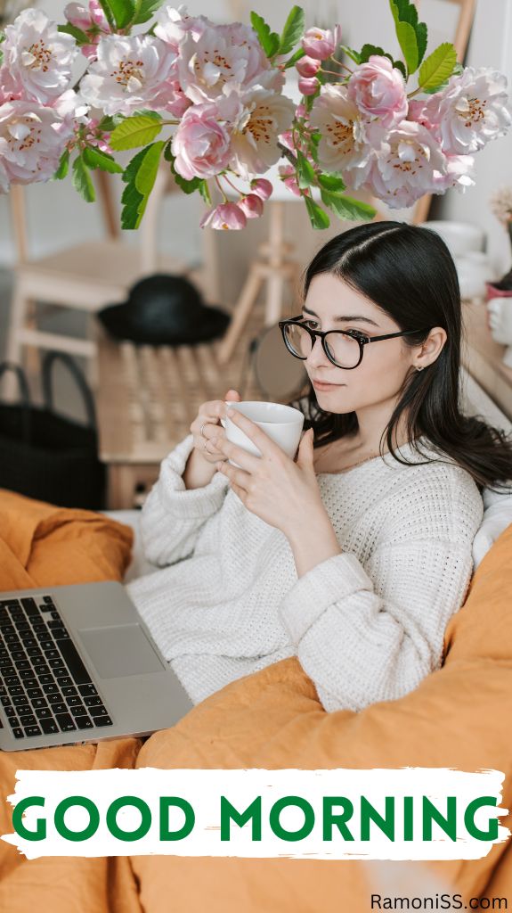 In the photo, a girl wearing a white sweater and glasses is sitting on her yellow bed, in whose hands the cup of tea and laptop are in her lap, and good morning is also written in the photo.