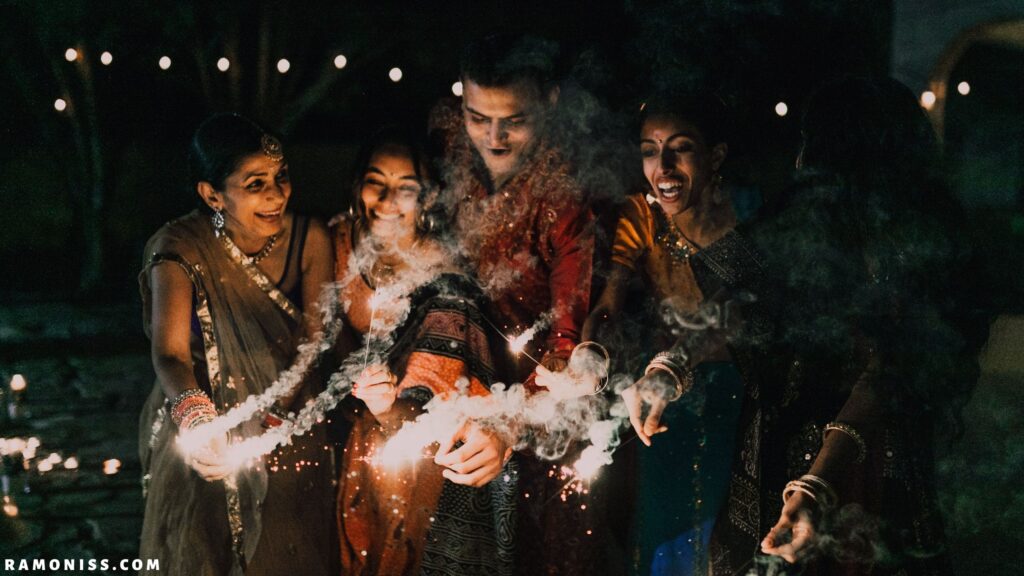 In the image, five members of a happy indian family including four ladies and a gentleman dressed in colorful clothes are lighting diwali sparklers at night.