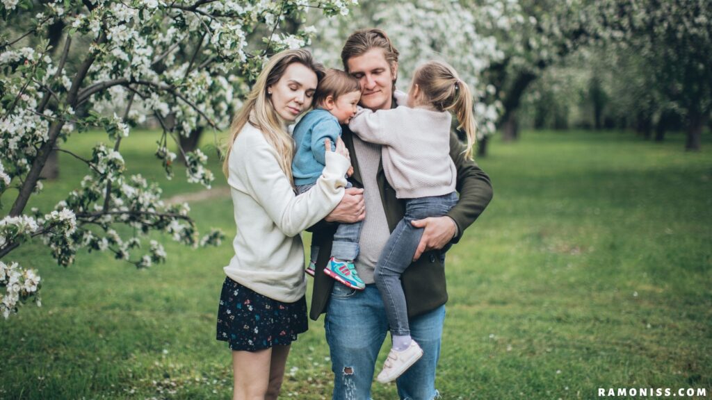 In the photo, two small children in the lap of the father standing in a garden and their mother standing together are showing love