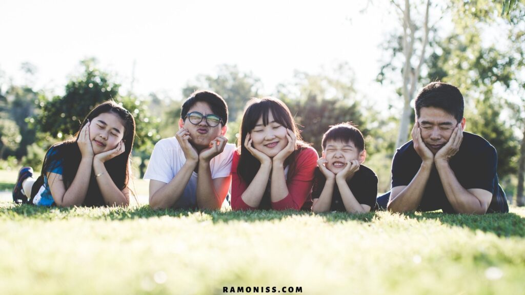 Five members of a happy family images are lying on the grass in a park and making faces like emoji with their hands on their chins.