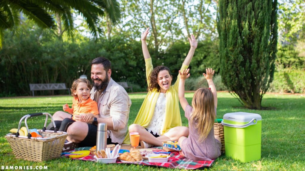 In the photo, a small child is sitting in the park with his father, mother and sister, all four people are celebrating a picnic and a sheet is spread on the ground, on which some food items are kept.