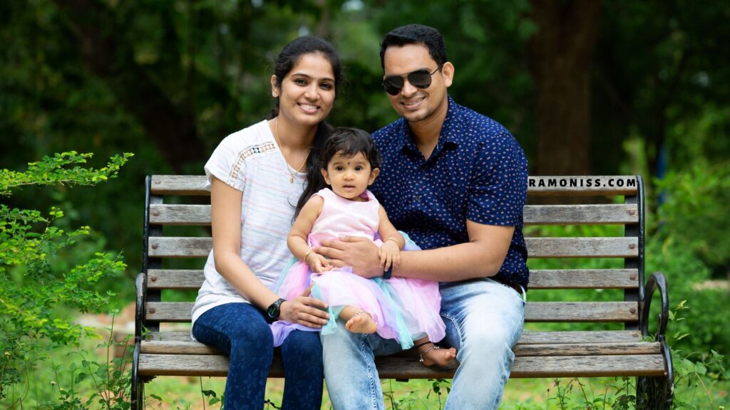 In the image, a happy mother and father sit on a park chair with their little girl in their arms.